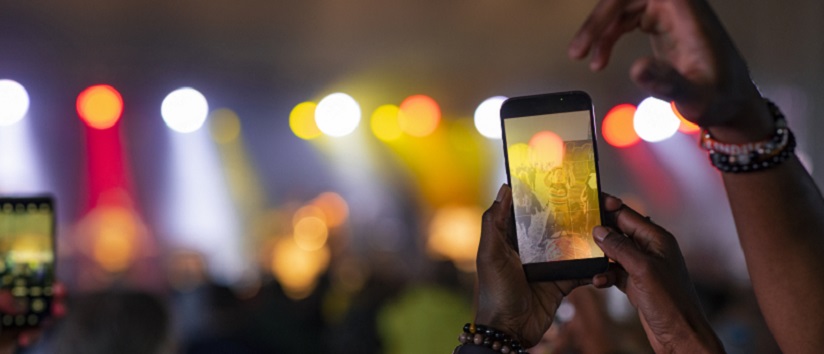 A point of view shot of an unrecognisable woman at a festival taking a picture on her smartphone of a live music festival. Everyone in the crowd is dancing together and having a good time. They are happy and sharing positive energy while watching the live show.