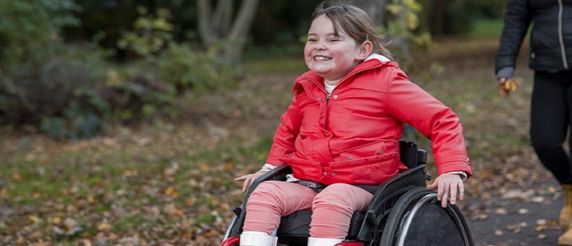 Young girl out with her family for a day out in a public park in Newcastle upon Tyne. She is a wheelchair user and is pushing herself ahead of her family in the park.