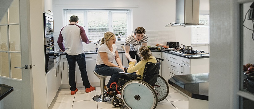 Mature mum and her son are interacting with her disabled daughter who is sitting in a wheelchair. The father is standing behind them preparing lunch.