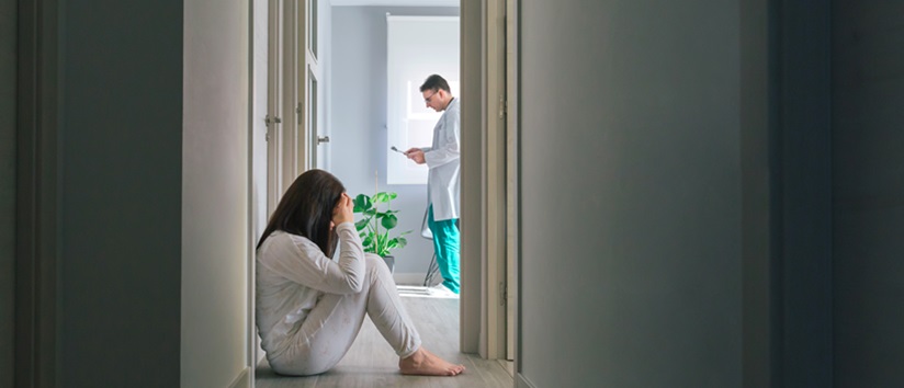 Female patient in pajamas sitting on the floor in the corridor of a mental health center with a doctor in the background