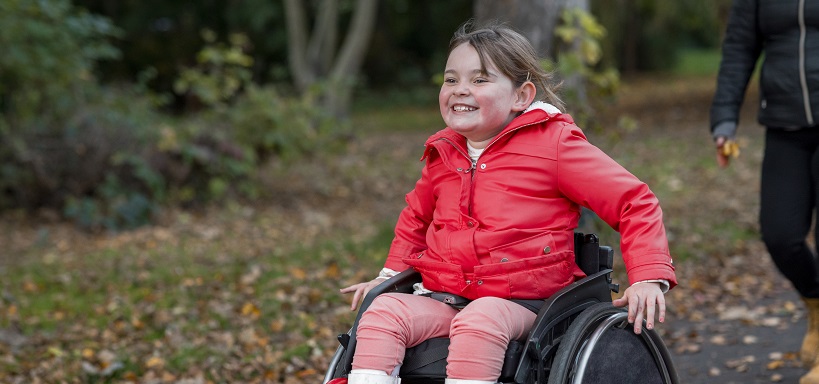 A low-angle view of a young girl out with her family for a day out in a public park in Newcastle upon Tyne. She is a wheelchair user and is pushing herself ahead of her family in the park.